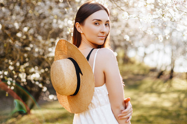 Girl with a straw hat in the spring in the park. Brunette with long hair holds a hat on a background of summer nature. Youth and beauty. - Fotografie, Obrázek