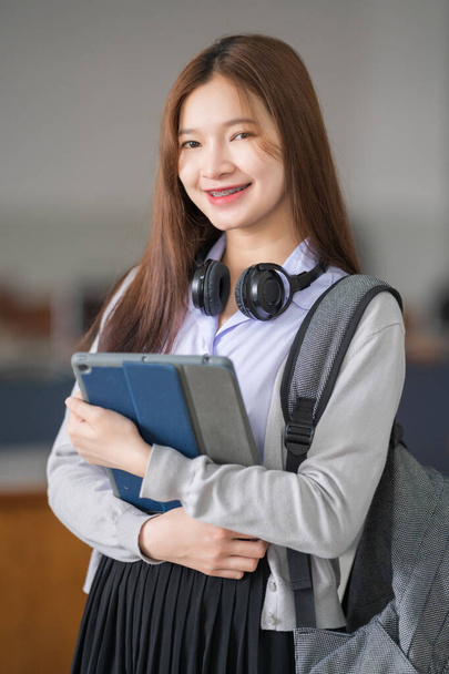 Portrait of young Asian woman student in uniform holding tablet in smart and happy pose in university or college classroom. Youth girl student and tutoring education with technology learning concept. - Photo, Image