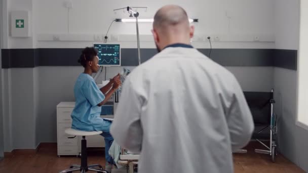 Передній вигляд хворої жінки лежить на ліжку, афро-американська медсестра аналізує рентгенівські кістки
 - Кадри, відео