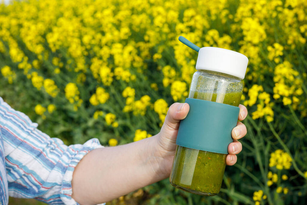  Eine Spaziergängerin hält eine Glasflasche mit vorbereitetem Obst und Gemüse in der Hand. Smoothie, köstliches grünes Getränk. Blühende gelbe Blumen im Hintergrund. Konzept, gesunde Lebensweise, Ernährung  - Foto, Bild