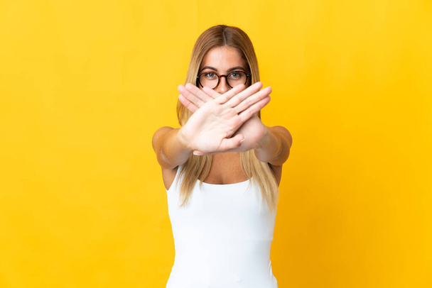 Mujer rubia joven aislada sobre fondo amarillo haciendo gesto de parada con la mano para detener un acto - Foto, Imagen