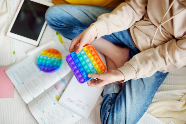 Teenage girl playing with rainbow pop-it fidget toy while studying at home. Teen kid with trendy stress and anxiety relief fidgeting game. Popping the dimples of sensory silicone toy. - Φωτογραφία, εικόνα