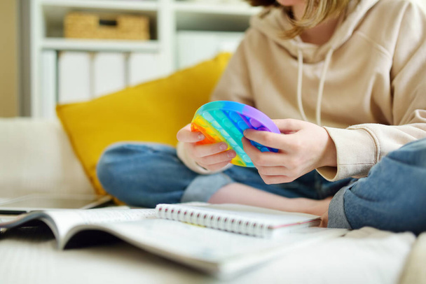 Teenage girl playing with rainbow pop-it fidget toy while studying at home. Teen kid with trendy stress and anxiety relief fidgeting game. Popping the dimples of sensory silicone toy. - 写真・画像