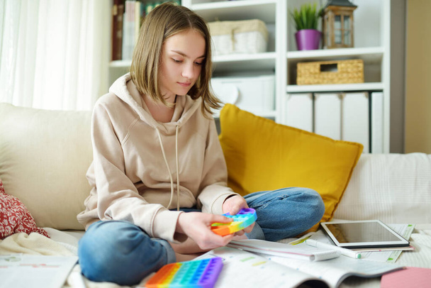 Teenage girl playing with rainbow pop-it fidget toy while studying at home. Teen kid with trendy stress and anxiety relief fidgeting game. Popping the dimples of sensory silicone toy. - Valokuva, kuva