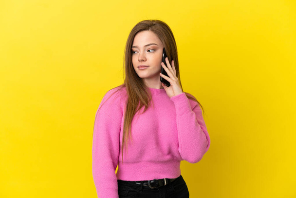 tiener meisje met behulp van mobiele telefoon over geïsoleerde gele achtergrond kijken naar de zijkant - Foto, afbeelding