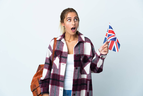 Młoda kobieta trzyma brytyjską flagę odizolowaną na białym tle, patrząc w górę i z zaskoczeniem - Zdjęcie, obraz