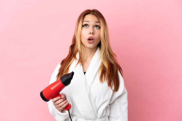 Teenager blonde girl holding a hairdryer over isolated pink background looking up and with surprised expression - Fotografie, Obrázek