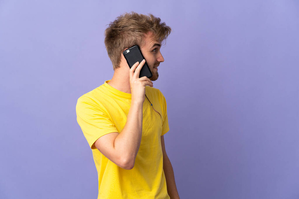 Fiatal jóképű szőke férfi elszigetelt lila háttér tartja a beszélgetést a mobiltelefon valakivel - Fotó, kép