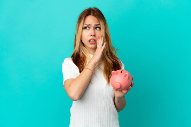 Teenager blonde girl holding a piggybank over isolated blue background whispering something with surprise gesture while looking to the side - Foto, Imagen