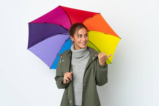Молодая женщина держит зонтик на белом фоне, указывая в сторону, чтобы представить продукт - Фото, изображение