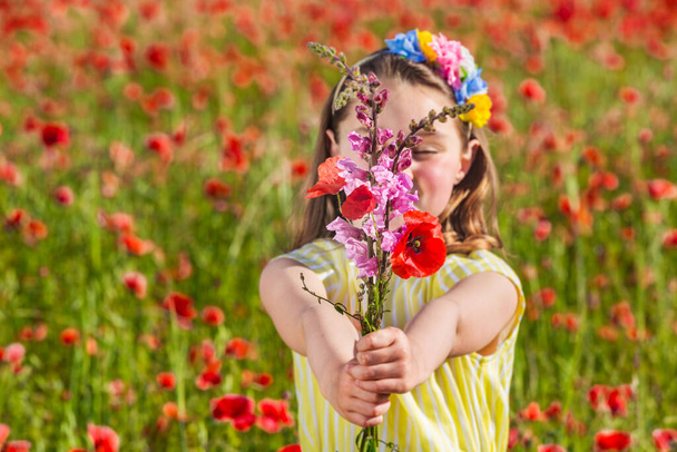 Cute little girl giving bouquet of fresh flowers to camera while resting in blooming field in summer day in countryside - Zdjęcie, obraz