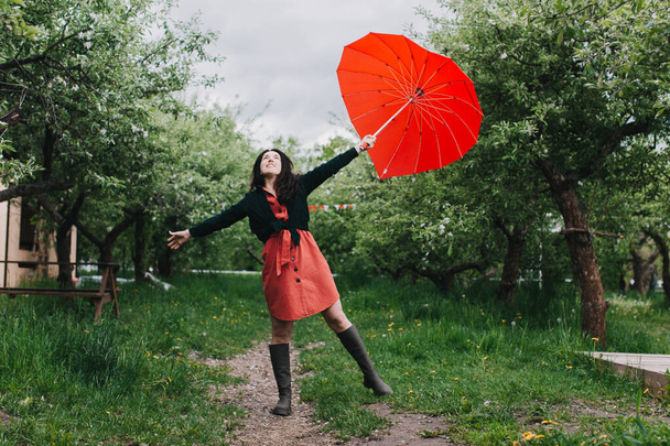 Όμορφη νεαρή γυναίκα κουβαλάει ομπρέλα σε σχήμα καρδιάς και κοιτάζει την κάμερα με χαμόγελο στον κήπο. Περνάμε καλά κάτω από τη βροχή - Φωτογραφία, εικόνα