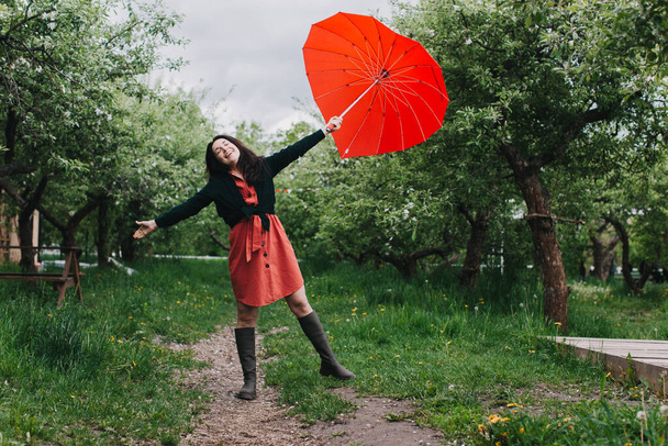 Kaunis nuori nainen kantaa sydämen muotoista sateenvarjoa ja katsoo kameraa hymyillen puutarhassa. Hauskaa sateen alla - Valokuva, kuva