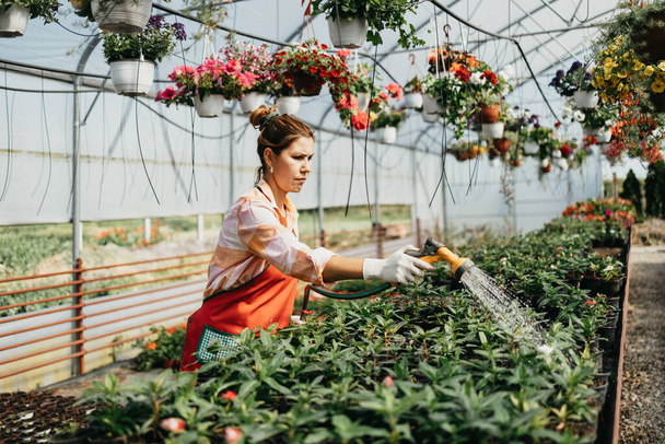 Happy and positive young adult woman working in greenhouse and enjoying in beautiful flowers. - Фото, зображення