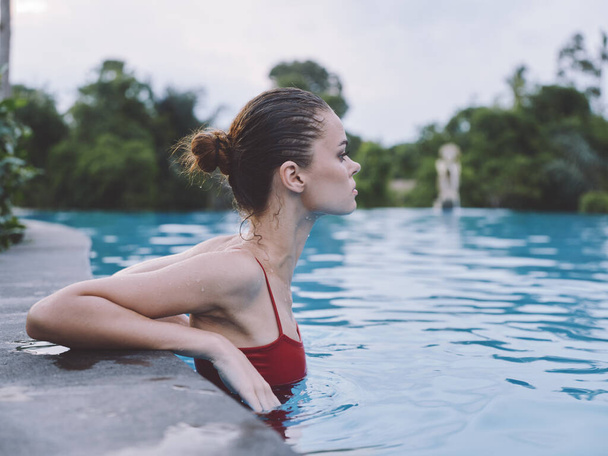 een vrouw in een badpak leunt op de tegels van het zwembad en staat in transparant water zijaanzicht - Foto, afbeelding
