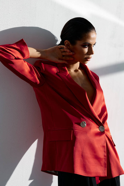 stijlvolle vrouw in een rode jas leunt tegen de muur binnen en kijkt naar de zijkant - Foto, afbeelding