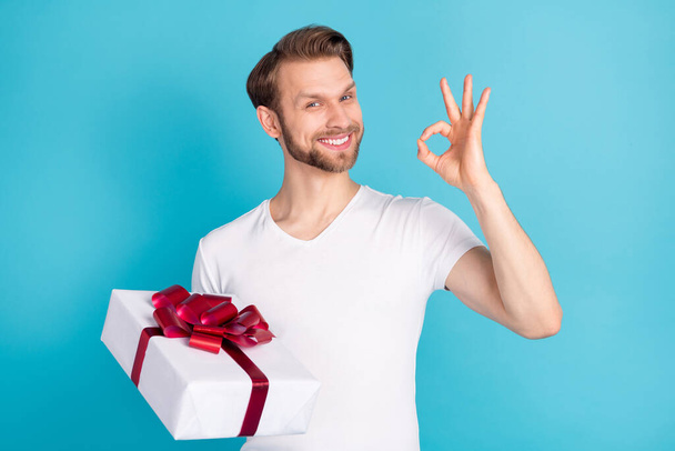 Photo of positive young man hold giftbox fingers show okey symbol isolated on blue color background - Foto, imagen