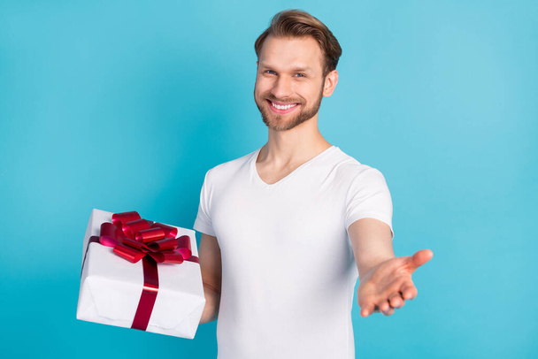 Photo of friendly candid man hold giftbox raise hand palm invite you toothy smile isolated on blue color background - Φωτογραφία, εικόνα