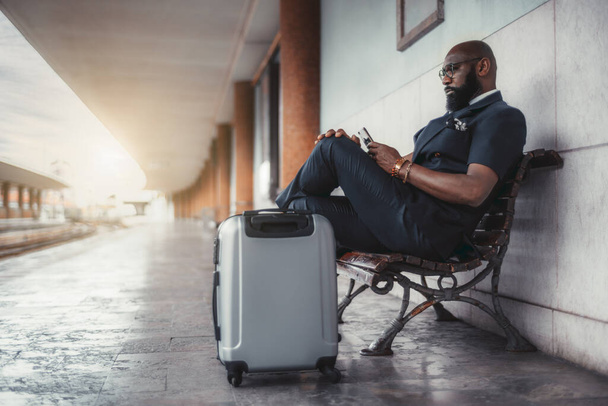 A serious black man entrepreneur on the railroad platform with his bags is phoning while waiting for a train; an African businessman with luggage on the railway station depot before his business trip - 写真・画像