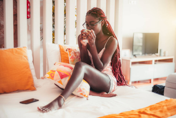 Morning tea of a dazzling sensual young sleepy African female with long braided afro hair and eyeglasses and nightie, she is drinking from the cup in her hands while sitting barefoot on her bed - Foto, Imagen