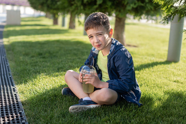Boy 7-8 years old drinks lemonade from a plastic glass in the park and looks at the camera - Фото, изображение
