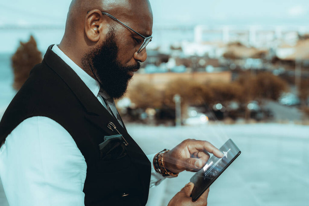 A side view of an elegant mature bald black guy entrepreneur outdoors in eyeglasses and fashionable business outfit, with a well-groomed beard, using a glowing user interface of a futuristic tablet pc - Фото, зображення