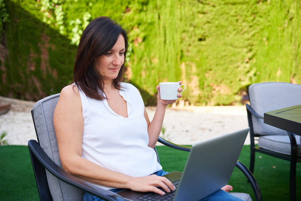 Una donna bruna caucasica tiene una tazza di caffè mentre lavora sul suo computer portatile - Foto, immagini