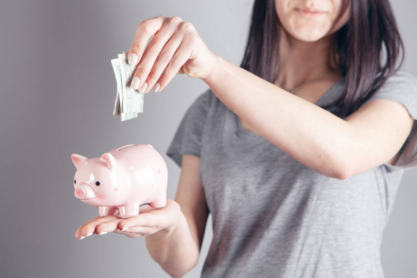 young woman throws dollar into piggy bank on gray background - Valokuva, kuva
