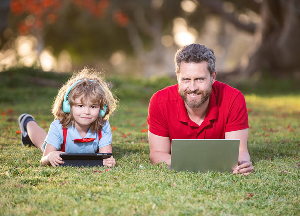 μπαμπάς και γιος χρησιμοποιούν φορητό υπολογιστή για βιντεοκλήση ή μάθημα ακούστε μουσική στα ακουστικά στο πάρκο, την οικογένεια. - Φωτογραφία, εικόνα
