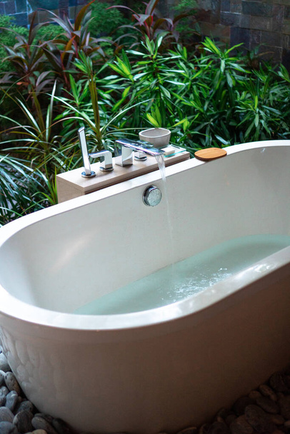 Moderne vrijstaande witte badkuip met stromend water, in de weelderige groene privétuin van het boetiekresort. Tropische vibes en sfeer van onberispelijke ontspanning. - Foto, afbeelding