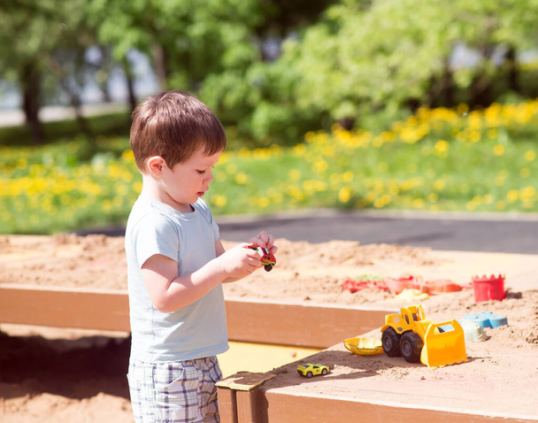 Un niño juega con juguetes, coches en la arena en el patio de recreo - Foto, Imagen