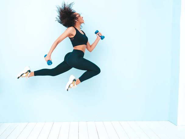 Fitness smiling black woman in sports clothing with afro curls hairstyle.She running and jogging . Young beautiful model with perfect tanned body.Female jumping with dumbbells in studio near blue wall - Foto, immagini