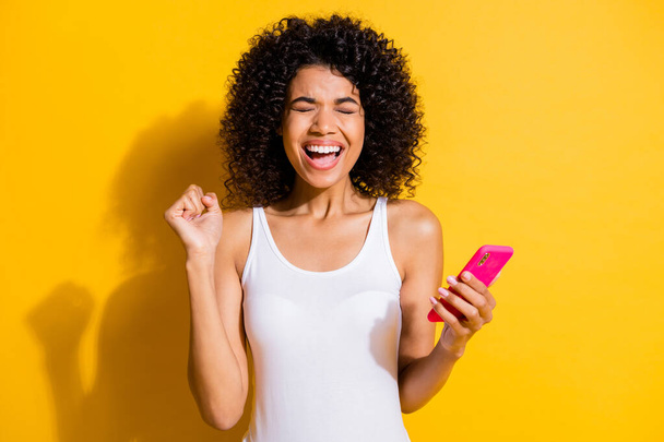 Photo of victorious afro american brunette girl raise fist hold phone wear white tank-top isolated on yellow color background - 写真・画像
