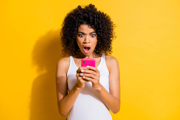 Photo of charming shocked young dark skin woman dressed white tank-top looking modern gadget isolated yellow color background - Fotografie, Obrázek