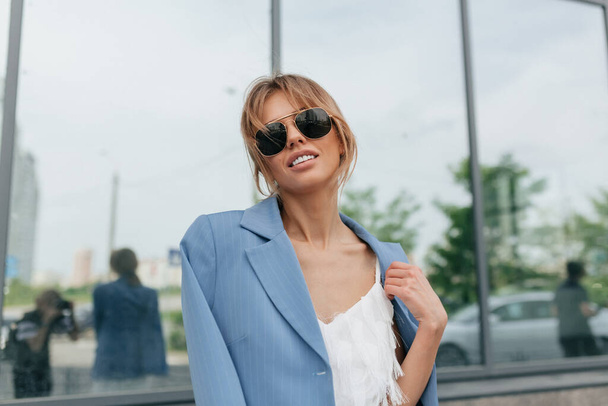 ガラスの建物を背景にカメラにポーズをとったブルーのジャケットとサングラスを身に着けているスタイリッシュな女性。高品質の写真 - 写真・画像