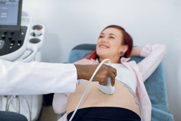 Cropped close up of a belly of happy cheerful pregnant woman having ultrasound scanning at the medical clinic. Female doctor making ultrasonic diagnostics. Focus on ultrasound transducer. - Foto, Imagem