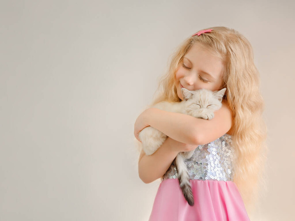 Küçük mutlu sarışın kız bej renkli İskoç bir kediyi kucaklıyor ve parlak bir elbiseyle gözlerini kapatarak gülümsüyor.. - Fotoğraf, Görsel
