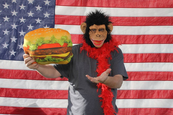 Человек носит маску обезьяны или шимпанзе, держа смешной реквизит подушки в фотобудке против американского флага. Фотобудки популярны на свадьбах и всех вечеринках и мероприятиях. - Фото, изображение