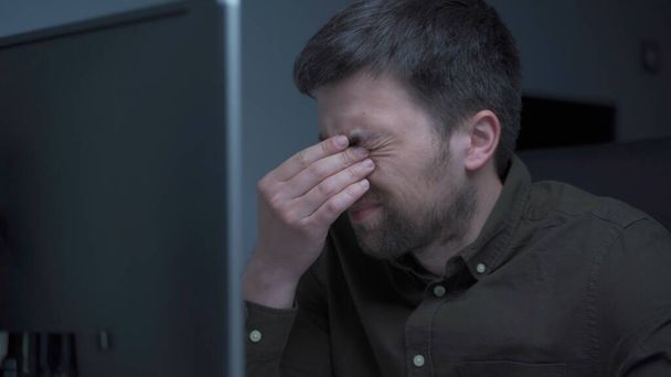 Valkoihoinen mies tekee ylitöitä tietokoneella ja silmien väsymistä kotitoimiston monitorista. Mies ottaa stressaavaa aikaa työskennellä PC. Uupunut ohjelmoija stressaavaa työtä, stressiä työpaikalla. - Valokuva, kuva