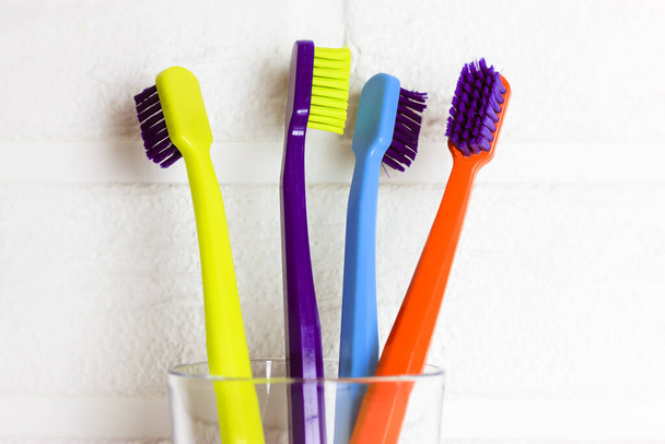 Set of plastic multicolored toothbrushes - green, blue, red, purple - in the transparent glass on a white background. Cup with four bright colorful soft toothbrushes against brick wall. Dental care.  - Photo, Image