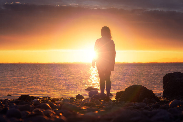 Adventure Caucasian Adult Woman Standing on a rocky beach on the West Coast Pacific Ocean watching a Dramatic Sunset. White Rock, Greater Vancouver, British Columbia, Canada. - Photo, image