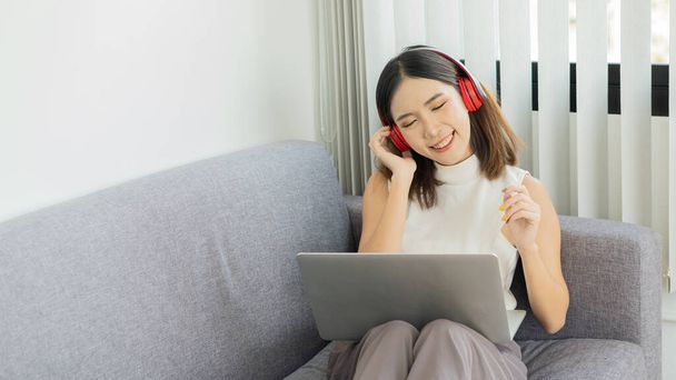 Een jonge vrouw die een film kijkt, naar muziek luistert op een laptop en thuis een hoofdtelefoon op een bank draagt. - Foto, afbeelding