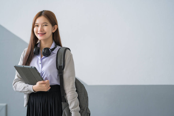 Portrait of young Asian woman student in uniform holding tablet in smart and happy pose in university or college classroom. Youth girl student and tutoring education with technology learning concept - Photo, Image