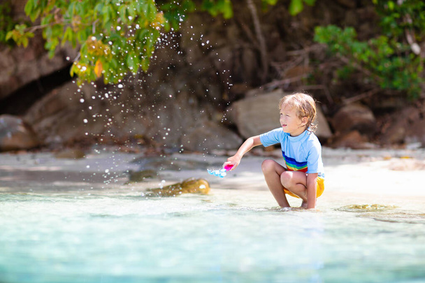 Niños jugando en la playa tropical. Agua salpicada de diversión. Un niño salpicando olas marinas. Los niños nadan y juegan en vacaciones familiares de verano. Juguete de arena y agua, protección solar para niños pequeños.  - Foto, imagen