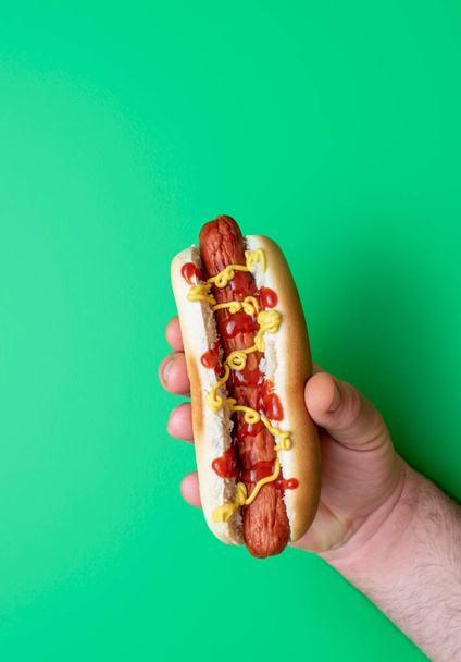 La mano di un uomo tiene un hot dog sopra un tavolo verde. Delizioso hot dog con senape e ketchup. - Foto, immagini