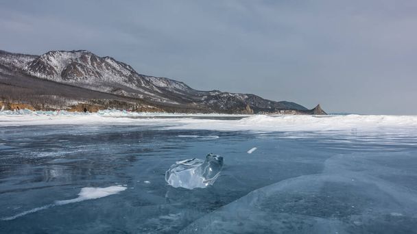 Outo jäälohkare on jäätyneellä järvellä. Häikäisy reunalla, heijastus. Syviä halkeamia, lunta jään pinnalla. Vuorijono taivasta vasten. Baikal - Valokuva, kuva