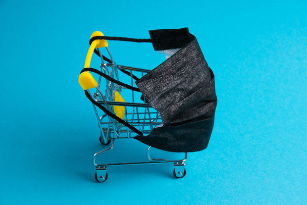 Medische masker en supermarkt trolley voor shopper op blauw papier achtergrond. Covid 19 concept quarantainebeperkingen. - Foto, afbeelding