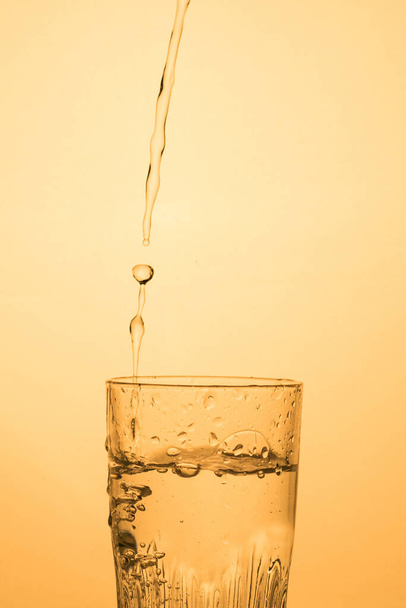 Una corriente de agua o vodka se vierte en un vaso con salpicaduras, sombra cálida, formato vertical con espacio para copiar. - Foto, Imagen