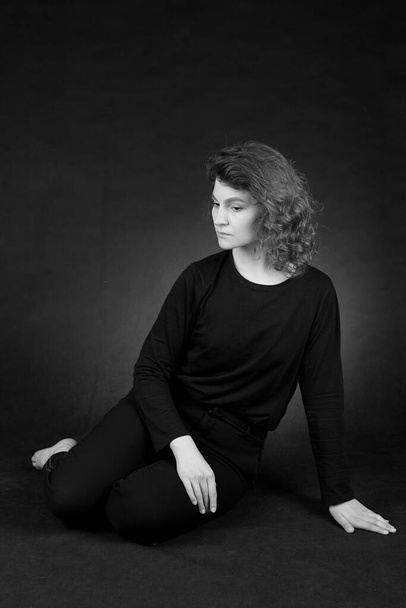 Portrait of a young attractive girl in black clothes on a dark background. The woman is sitting on the floor. Black and white photography. - Фото, зображення