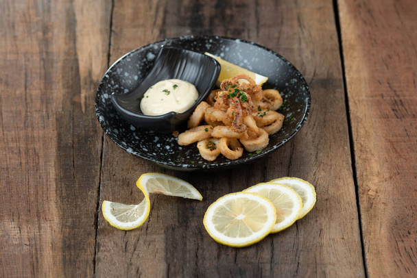 Un plato de crujientes anillos de calamares fritos, calamares con pimiento rojo ahumado, almendras, vinagre de cereza, tomate y cayena servido con salsa tártara inmersión en el lado aislado en mesa de madera - Foto, imagen